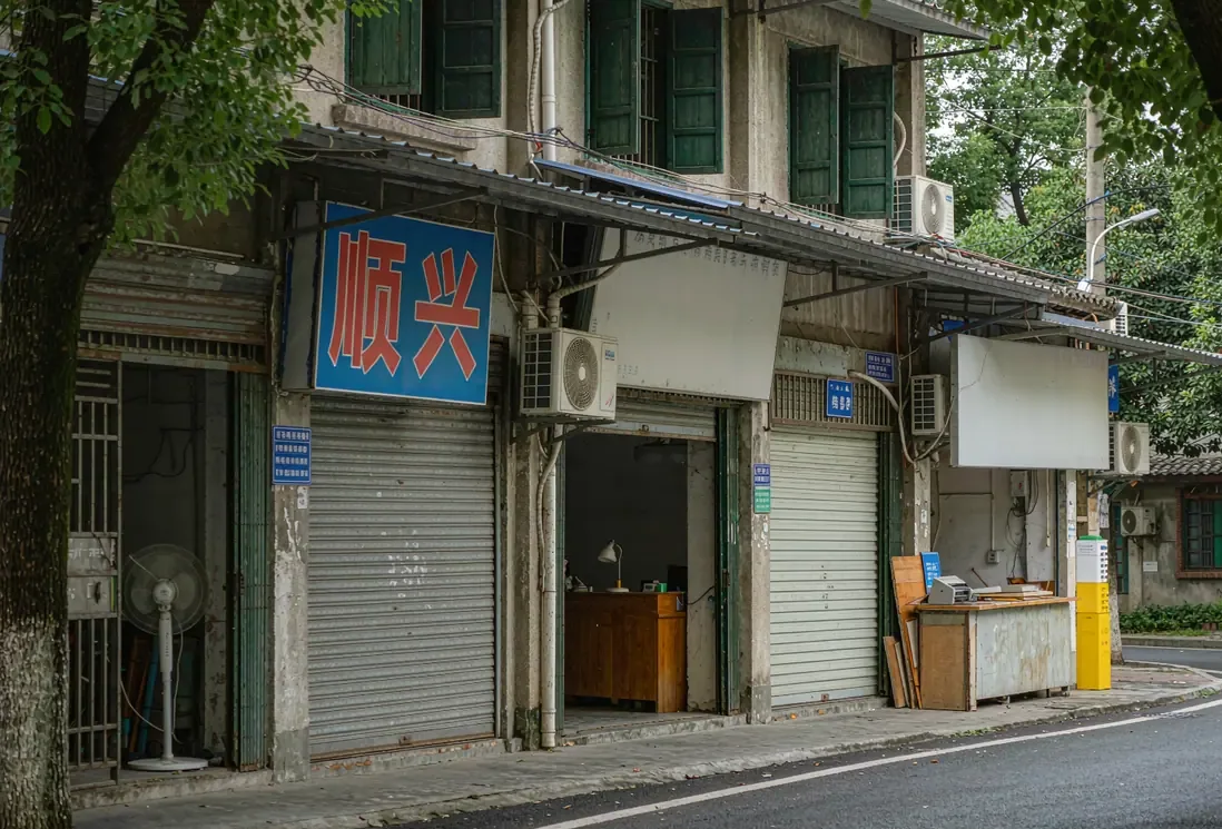 南方小镇街边三家广告小门市并排,柜台后摆着小型喷绘设备,阴天自然光(AI生成示意图)